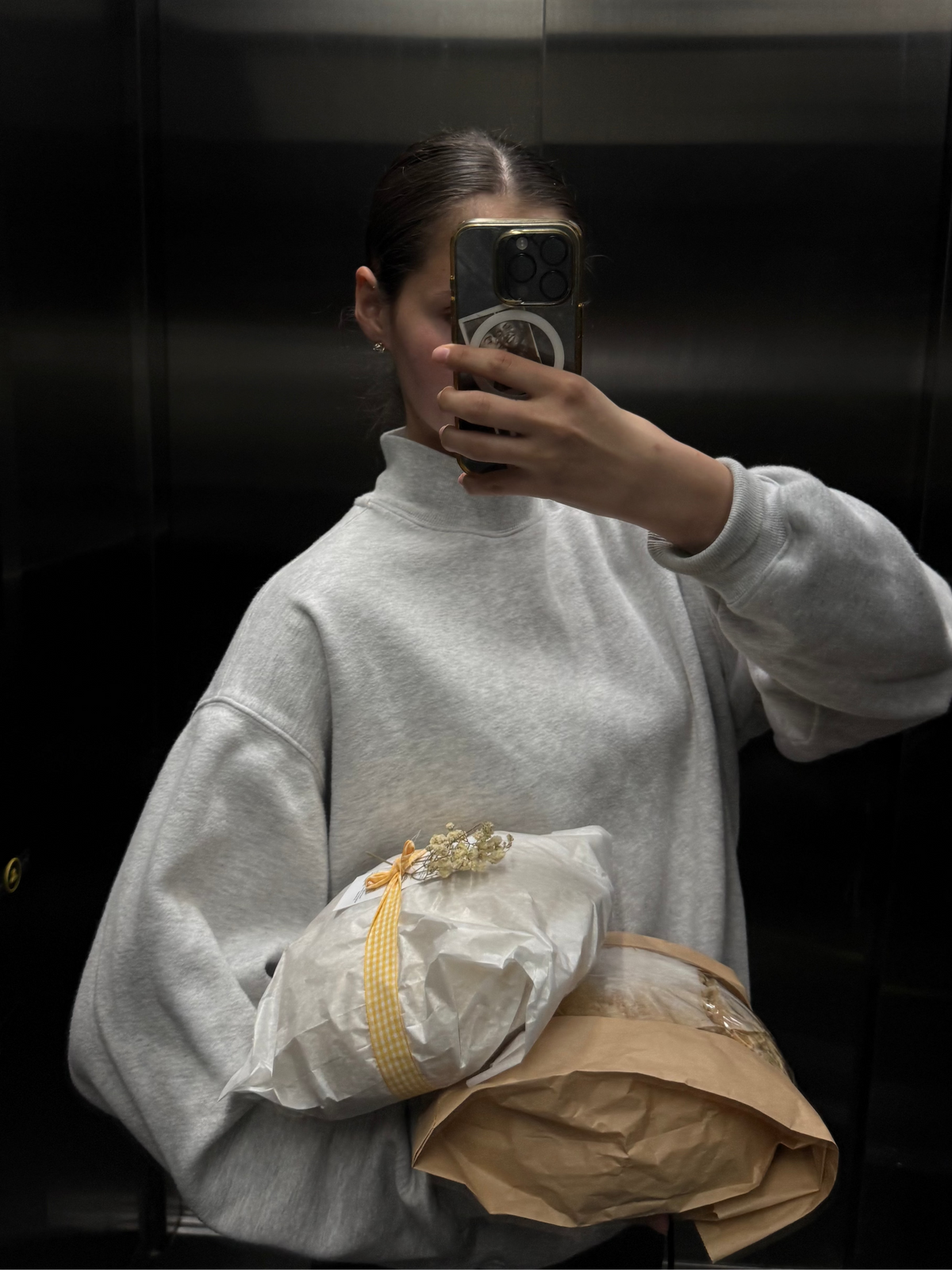 Person taking a mirror selfie wearing a light gray sweater, holding a wrapped package.